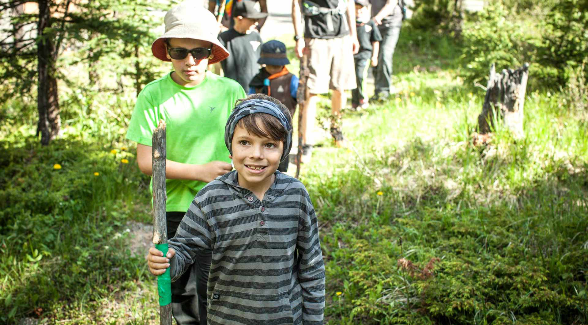 A group of young scouts hiking a trail through a forested area, and the lead person is holding a walking stick.