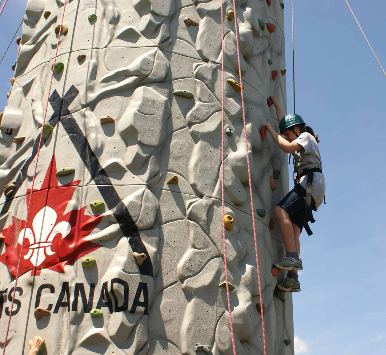 A Scout in harness and safety helmet climbing a cement vertical climbing wall.