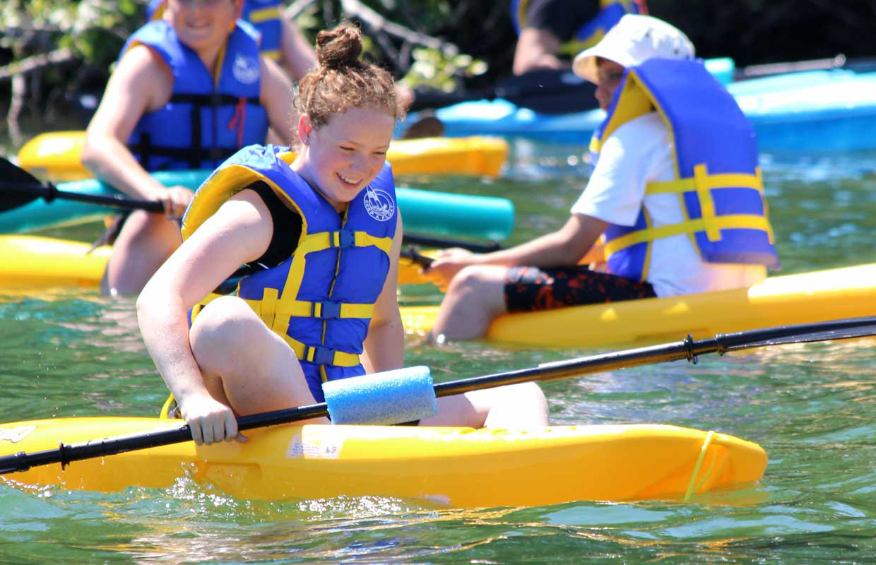 A group of young children wearing life vests on the water, in kayaks and paddling on a bright summer day.