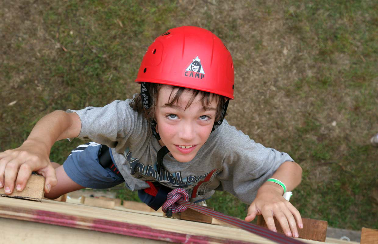 A young boy in a harness and safety helmet is looking up as he makes his way up a climbing wall.