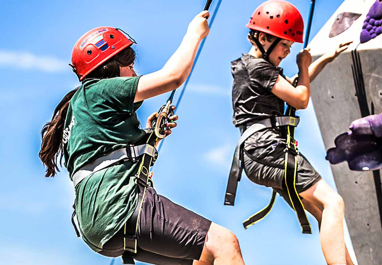 Two Scouts in harness and safety helmet climbing to the top of a cement climbing wall.