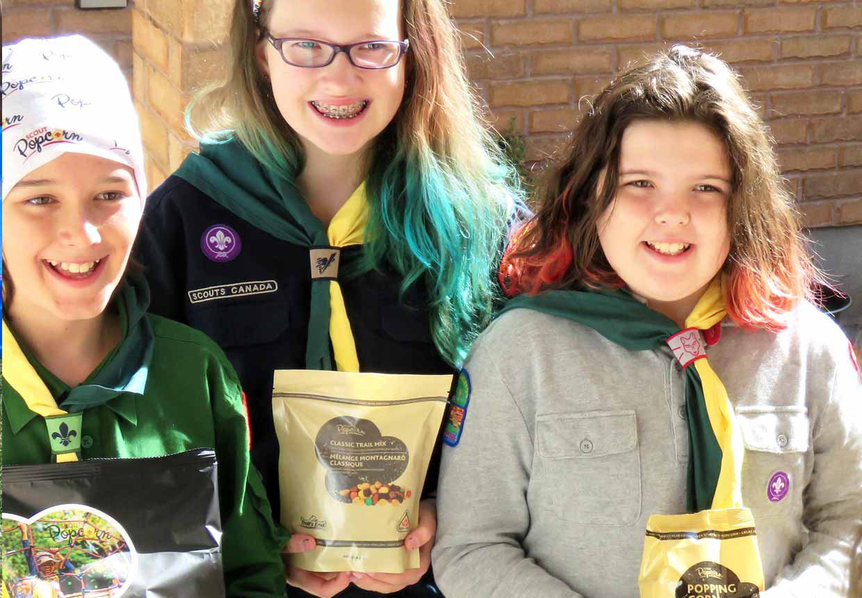 Three girl Scouts smiling and holding the popcorn and trail mix bags they are selling.
