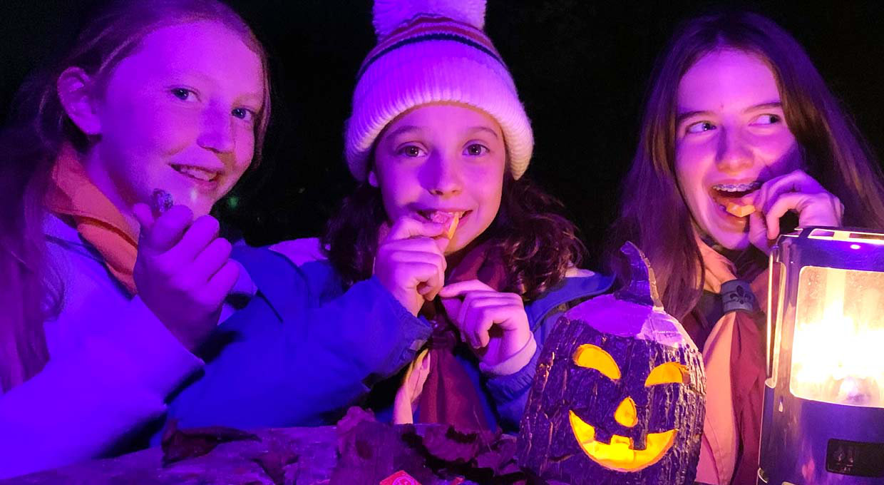 Three Girl Scouts standing at night with a lighted Halloween pumpkin eating snacks.