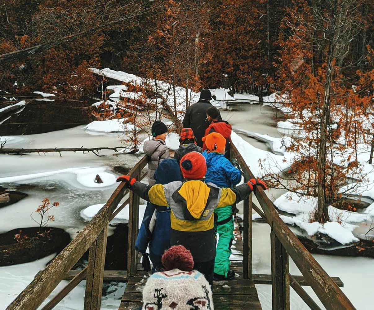 A group of Scouts is crossing a bridge over a small creek in the forest during a winter hike.