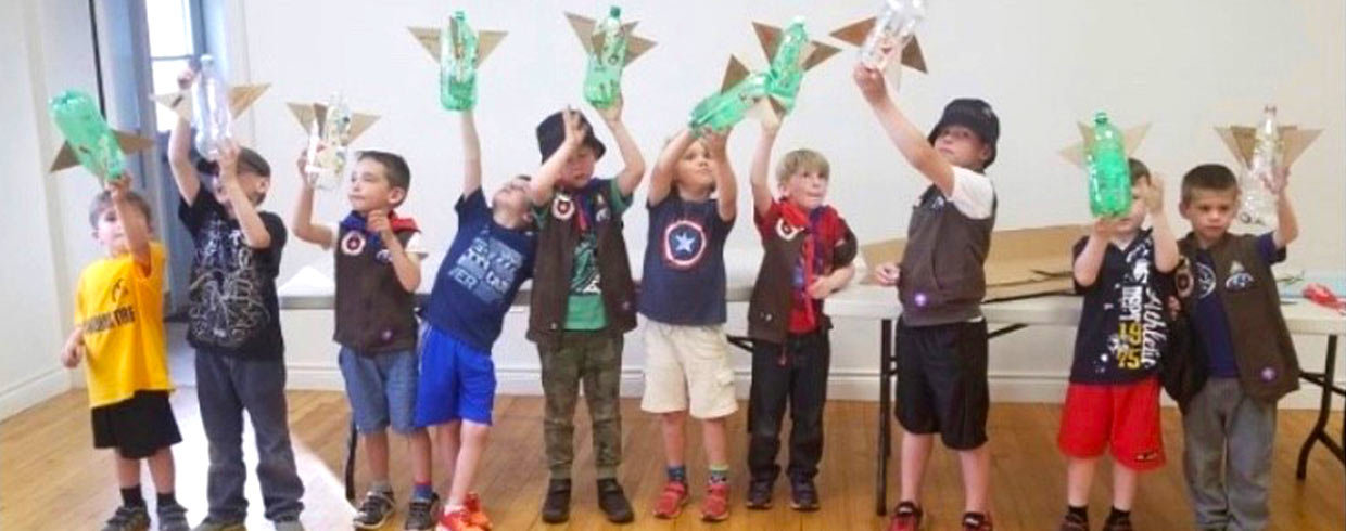 A group of young Scouts inside a craft room holding up their creations made from plastic bottles and cardboard.