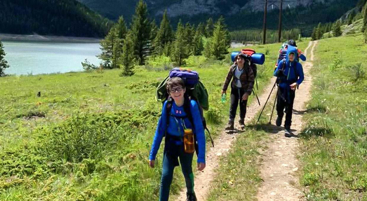 Three Venturers loaded down with full backpacks hiking along a dirt trail, surrounded by mountains and water.