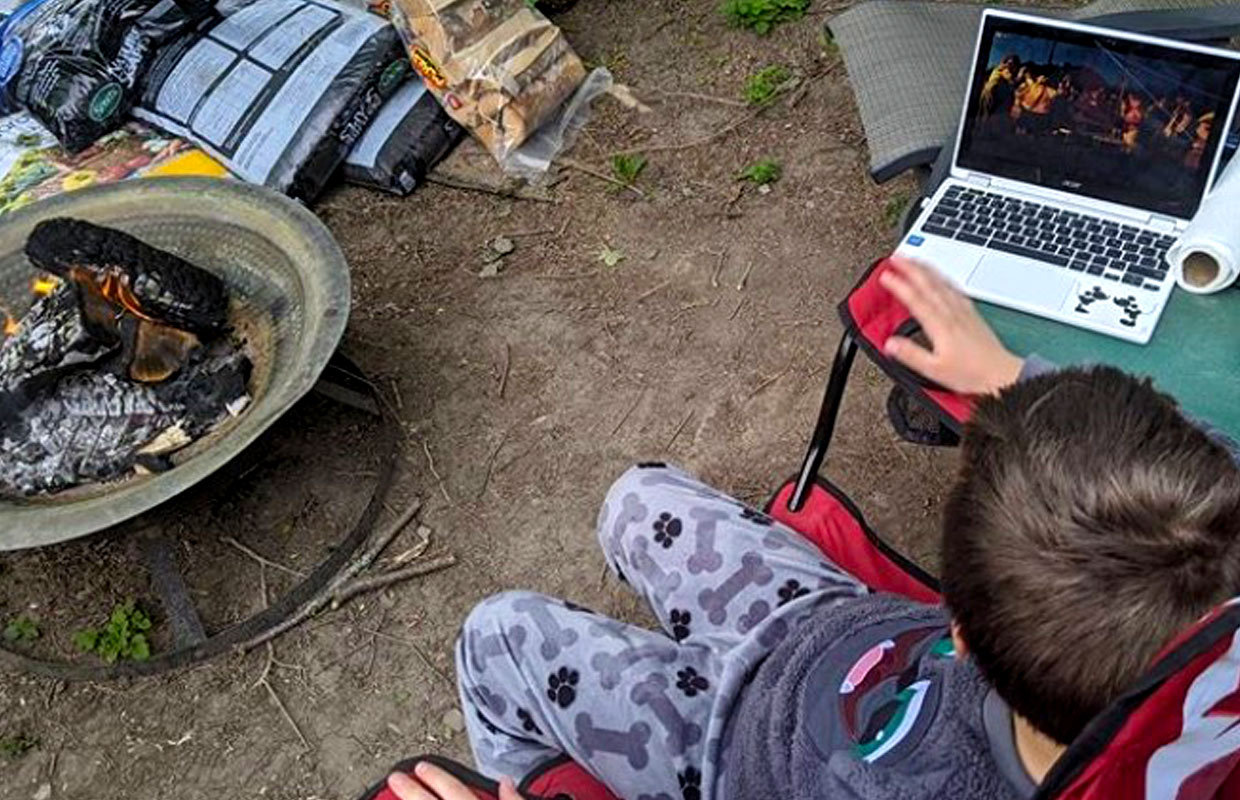 A young Scout watching virtual campfire activites on his laptop while sitting beside a live backyard fire.