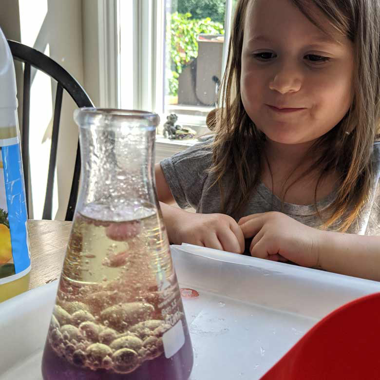 A smiling young girl Scout watching a beaker bubbling with two kinds of red and yellow liquids.