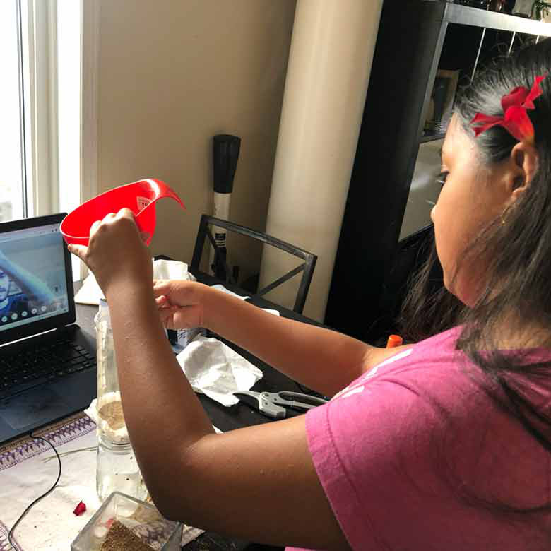 A young girl Scout creating a fun experiment using a funnel and glass jar, while watching instruction from her laptop.
