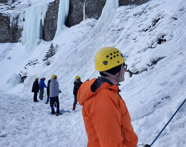 A group of Rovers and Venturers in harness and safety helmets preparing to ice climb a steep rocky cliff.