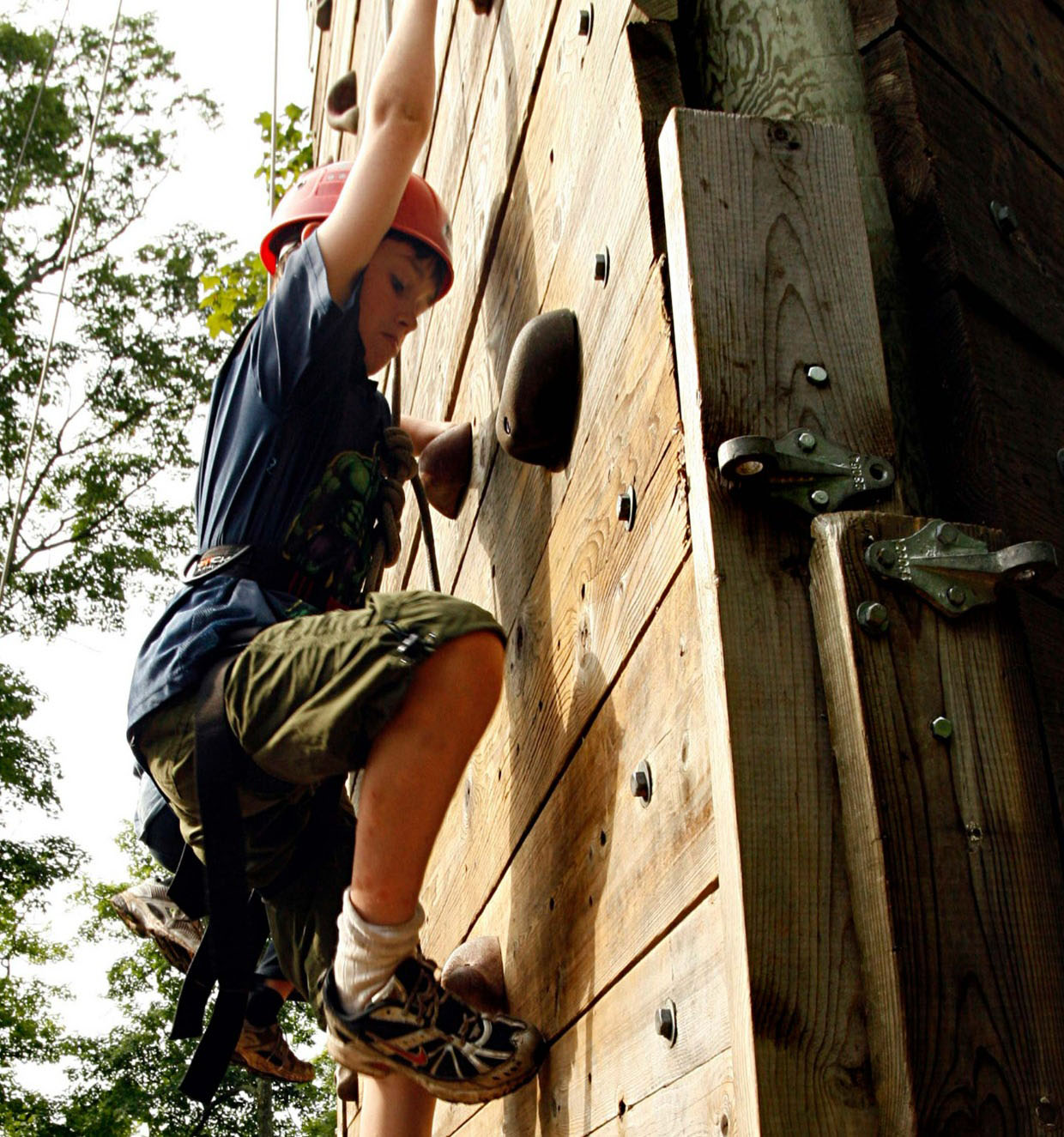 A boy scout wearing a safety helmet climbing to the top of a climbing wall.